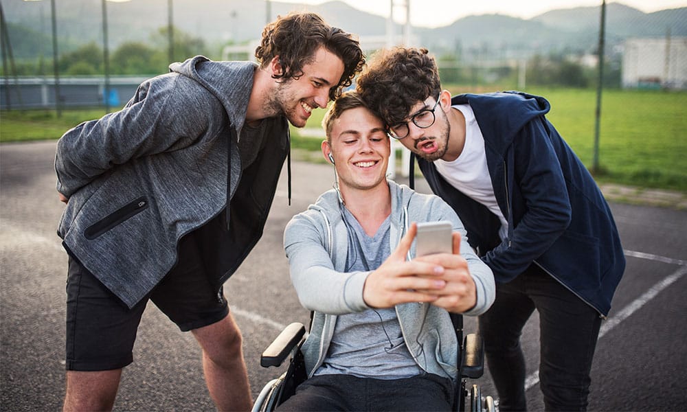 A boy in wheelchair with smartphone and teenager friends taking selfie on a playing field.