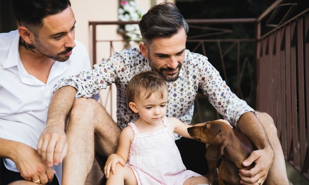 Gay couple with daughter and dog on balcony using digital tablet