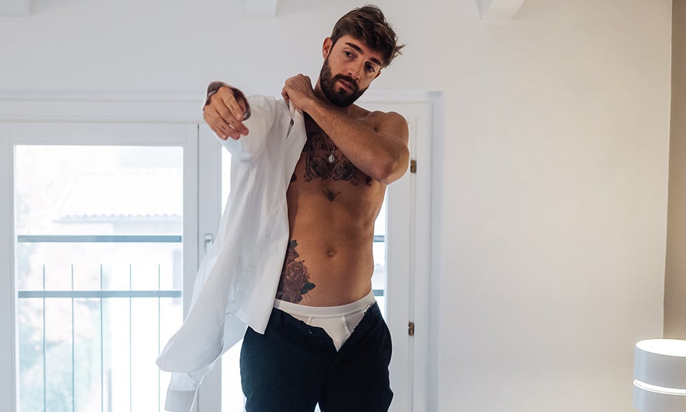 Adult man with tattoos putting on shirt in bedroom