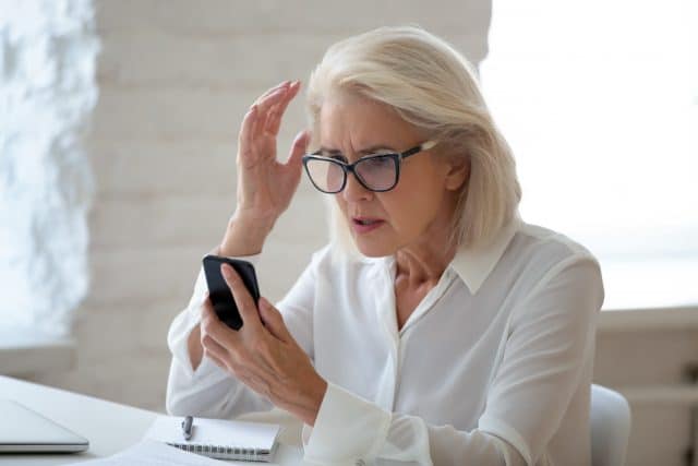 Confused senior businesswoman sit at office desk hold cellphone