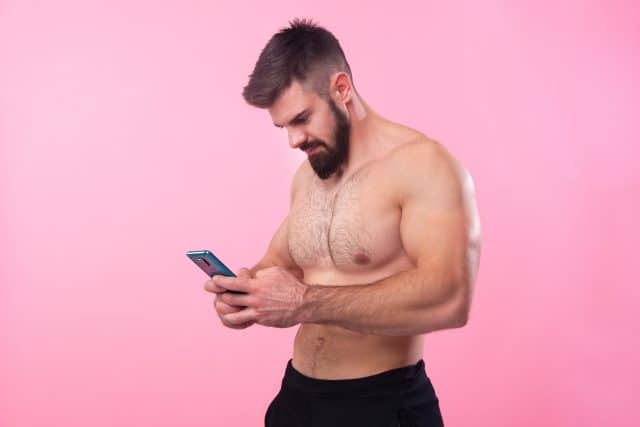 Shirtless strong bodybuilder talking on his cell, using a smartphone, pink studio background