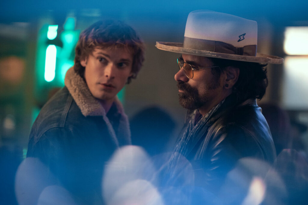 "Two men in a dimly lit indoor setting, one in a jean jacket with a fur collar looking back, the other with a hat and glasses facing forward, colorful lights blurred in the foreground."